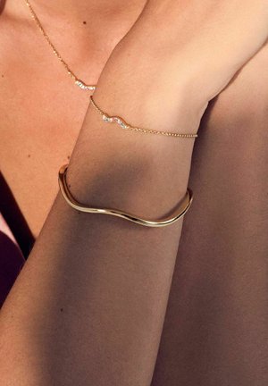 Guldarmband med vågig design, slät yta, i kombination med ett fint kedjearmband med flerfärgade ädelstenar.