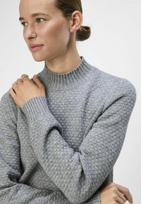 Suéter de punto gris texturizado con un alto cuello acanalado y mangas largas, que presenta un patrón de puntos en relieve y puños ajustados.