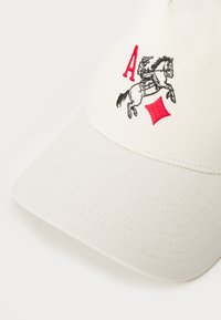 Hvit caps med bøyd skjerm, med svart og rød brodert design av en rytter på en hest og en rød diamantform.