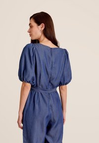 Denim jumpsuit met korte pofmouwen, een getailleerde taille en een V-hals. De stof heeft een middelblauwe tint met een gladde textuur. Achterkant met ritsdetail.