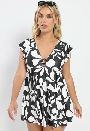 Mujer rubia con vestido corto de manga corta de flores en blanco y negro con detalle de anillo en el escote, pendientes plateados y gafas de sol en la cabeza.