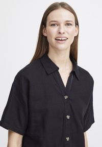 Camicia nera con bottone, colletto, maniche corte e vestibilità comoda; presenta bottoni arrotondati e una trama liscia.