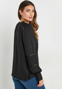 Blusa nera con motivo a pois testurizzati, scollatura arricciata e maniche lunghe con dettagli in pizzo. Tessuto morbido e vestibilità rilassata.