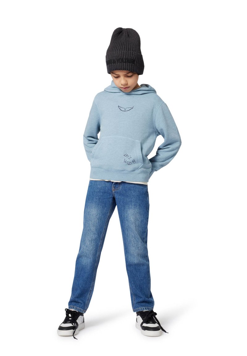 Kind met een zwarte beanie, lichtblauwe hoodie met de tekst "Laten we wild gaan", blauwe jeans en zwart-witte sneakers, staand met zijn handen achter zijn rug.