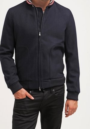 Mężczyzna ubrany w granatową kurtkę na zamek z prążkowanymi mankietami i dołem, z czerwono-białym paskowanym kołnierzem oraz czarne spodnie.