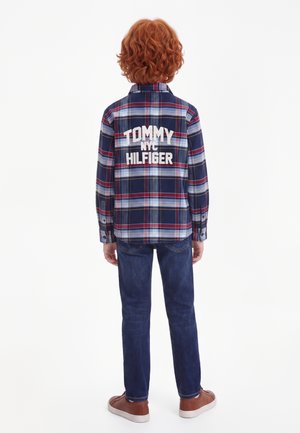 Kind met rood krullend haar, gekleed in een blauw- en rood geruit overhemd met de tekst "Tommy NYC Hilfiger" op de rug, blauwe spijkerbroek en bruine schoenen.