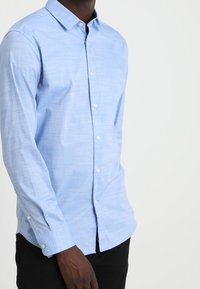 Ljusblå långärmad skjorta med krage som knäpps, med en subtil textur och vita knappar, tillsammans med svarta byxor.