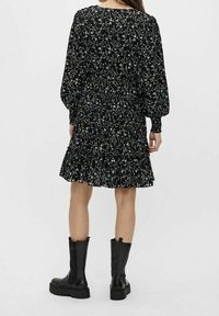 Robe noire à fleurs avec manches longues, taille smockée, ourlet à volants et motif de petites fleurs blanches et vertes. Associée à des bottes noires.