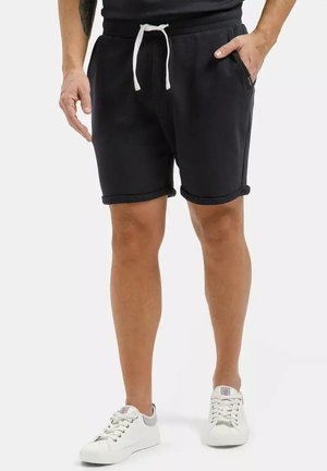 Mężczyzna w czarnych spodenkach ze sznurkiem i białych trampkach, stoi z jedną ręką w kieszeni na jednolitym tle.