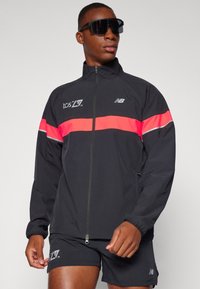 Black zip-up jacket with a bright pink horizontal stripe and reflective details. Made of lightweight material, featuring a high collar and cuffed sleeves.
