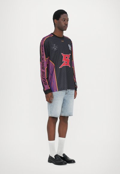 Langärmliges schwarzes Jersey mit lila und orangen Akzenten, das Grafiken und Text zeigt. Getragen mit hellblauen Jeansshorts und schwarzen Slippern.