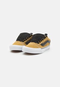Adidași din suede galben cu șireturi negre, talpă din cauciuc alb și o bandă laterală neagră. Prezentând cusături contrastante și o etichetă pe călcâi.