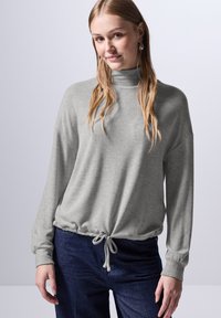 Lichtgrijs langemouwen shirt met een hoge hals en een verzamelde taille met trekkoord, gecombineerd met donkerblauwe jeans. Zachte stof met een ontspannen pasvorm.
