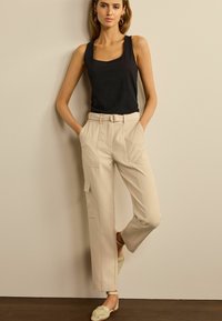 Zwarte mouwloze top gecombineerd met beige hoge taille broek met zijzakken en een riem. Model staat met haar handen in de zakken.