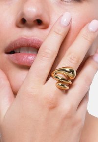 Anello d'oro con un design astratto e fluido composto da tre sezioni arrotondate collegate; superficie lucida e texture liscia. Il dito poggia vicino alle labbra.