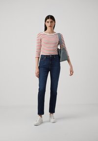 Stripet langermet topp i hvit og rød, kombinert med mørkeblå jeans og hvite sneakers. Modellen holder en lyseblå håndveske.
