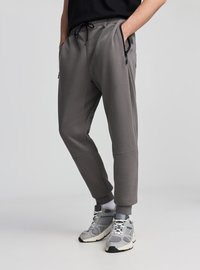 Szare spodnie dresowe z regulowanym sznurkiem w pasie, bocznymi kieszeniami na suwak, zwężanym krojem i ściągaczami na końcach nogawek, zestawione z szarymi sneakersami i białymi skarpetkami.