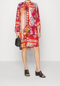 Monivärinen mekko, jossa on rohkeita kukka- ja geometrisia kuvioita, pitkät hihat, rypytetty kaula-aukko ja löysä istuvuus. Yhdistetty mustiin avokasneuloihin.