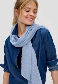 Écharpe bleu clair avec un motif texturé et en relief drapée autour du cou, associée à une chemise en jean sombre. Tissu doux et léger.