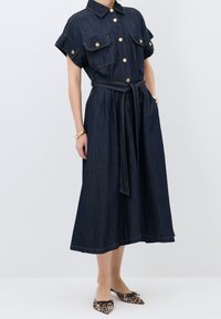 Robe en denim foncé avec des manches courtes roulées, des boutons dorés, deux poches poitrine, une ceinture à la taille et une jupe évasée. Portée avec des chaussures à imprimé léopard.