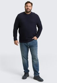 Navyblauwe gebreide trui met een textuurpatroon, gecombineerd met lichtblauwe denim jeans en zwarte sneakers. Volwassen mannelijke model poseert met handen in de zakken.