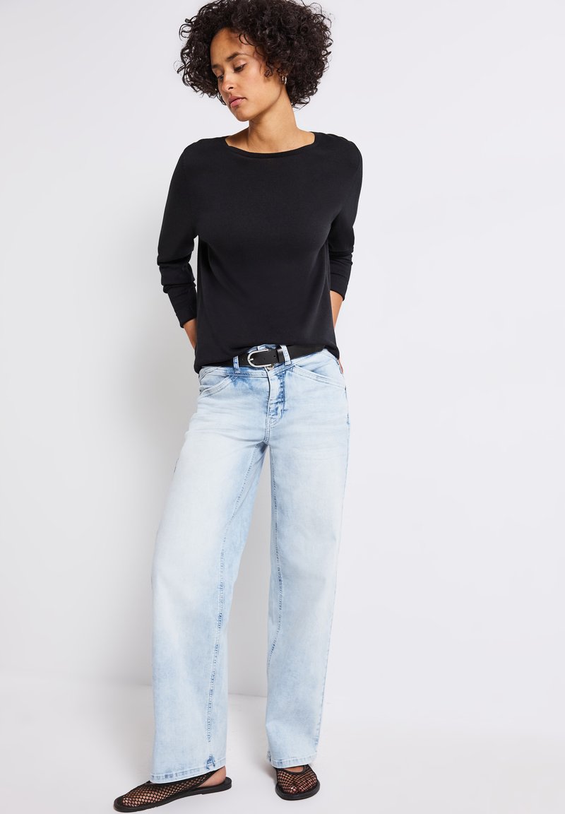 Svart långärmad topp, ljusblå vida jeans och svarta nät slip-on skor. Funktioner inkluderar en rund halslinje och bälte.