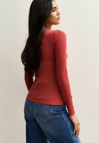 Femme aux longs cheveux noirs, portant un haut ajusté à manches longues couleur rouille et un jean bleu, se tenant de côté contre un mur clair.