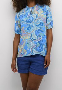Blouse bleu à motif paisley avec des manches courtes à volants et un décolleté froncé, associée à un short bleu. Le tissu semble léger et texturé.
