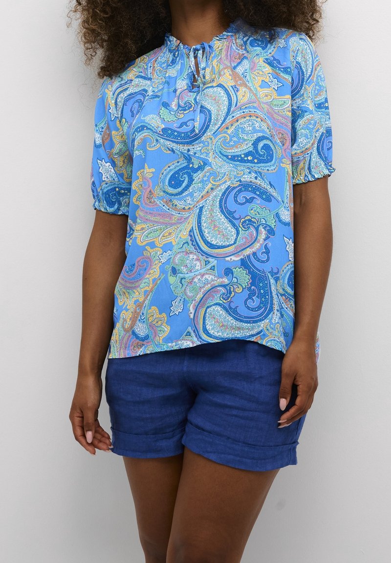 Blouse bleu à motif paisley avec des manches courtes à volants et un décolleté froncé, associée à un short bleu. Le tissu semble léger et texturé.