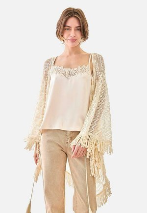 Mujer con camisola beige con ribete de encaje, cárdigan con flecos de crochet y pantalones marrón claro, de pie contra un fondo blanco liso.