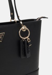 Musta olkalaukku, joka on valmistettu teksturoidusta synteettisestä materiaalista, ja siinä on kullanväriset yksityiskohdat, avainrenkaalla varustettu koriste sekä kaksi mustaa hintalappua, joissa on kohokuvioidut numerot.