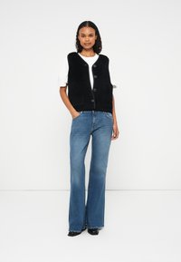 Black fuzzy vest with a button closure over a white shirt, paired with flared blue jeans and black shoes. Smooth texture and casual design.