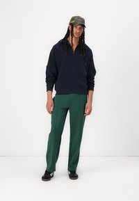 Marineblå hettegenser med halv glidelås, kombinert med grønne bukser med skreddersydd passform. Sorte joggesko og en camo-kapas fullfører antrekket.