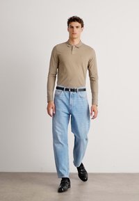 Beige színű, hosszú ujjú pólóing gallérral, világoskék bő szabású farmernadrág, fekete öv és fekete loaferek, világos háttér előtt sétálva.