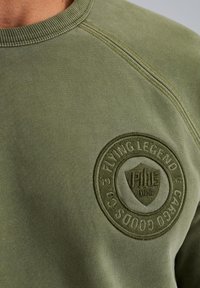 Olive green sweatshirt with a ribbed crew neckline, featuring a stitched round badge on the chest with embossed text and shield design.