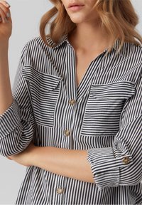 Chemise à rayures bleu marine et blanche. Comprend deux poches poitrine, des manches retroussées et des boutons ronds avec une texture lisse.