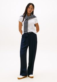 Sieviete stāv, tērpusies baltā T-kreklā, tumši zilās plašajās biksēs, baltos apavos un tumši zilā ar baltām svītrām džemperī, kas uzmests pār pleciem.