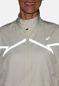 Vrouw die glimlacht in een lichtbeige ritsjack met witte reflecterende zigzagstrepen en een Asics-logo op de borst.