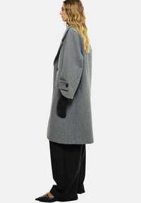 Manteau en laine gris à coupe loose, avec de gros boutons et de larges revers. Associé à des gants noirs et un pantalon noir large. Chaussures plates noires.