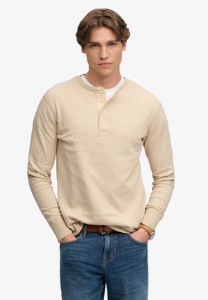 Jonge man met lichtbruin haar, die een beige henley met lange mouwen draagt, blauwe spijkerbroek, bruine riem en een polshorloge, met handen in de zakken.