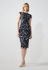 Abito nero drappeggiato con motivo astratto bianco, design aderente, lunghezza al ginocchio e maniche a cappuccio. Abbinato a sandali con tacco chiari.