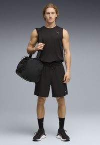 Μυώδης νέος άνδρας με μαύρο αμάνικο πουκάμισο, σορτς και αθλητικά παπούτσια, κρατώντας μια μαύρη αθλητική τσάντα στον ώμο, στέκεται μπροστά σε γκρι φόντο.