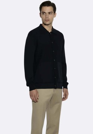 Uomo che indossa un cardigan nero con bottoni e pantaloni beige, in piedi con le mani rilassate ai lati, guarda leggermente a sinistra.