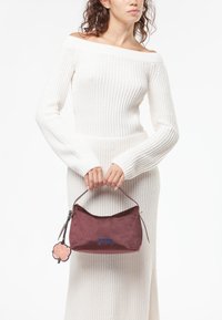 Diep bordeaux kleurige suède handtas met een gebogen bovenkant, voorzien van blauwe borduursels en een bloemen sleutelhangertje. Gladde textuur, compact ontwerp en ritsdetail.