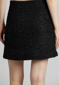 Falda de tweed negra con una superficie texturizada, forma acampanada y un dobladillo sin terminar. La vista trasera muestra un ligero brillo y detalles desgastados a lo largo de los bordes.