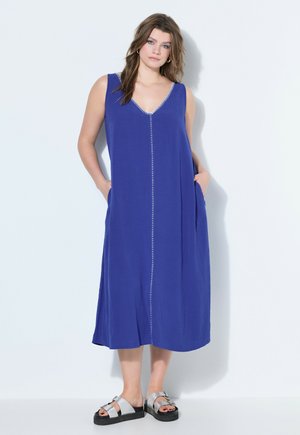 Frau trägt ein ärmelloses midi-kleid in königsblau mit vertikalen nähten vorne, steht mit händen in den taschen und silbernen sandalen.