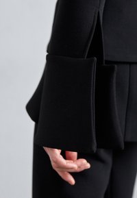 Gros plan d'une personne portant une veste noire à manches longues avec des poignets larges et surdimensionnés qui pendent librement autour du poignet.