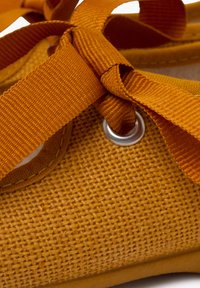 Žlutá plátěná bota s texturovaným povrchem, se zaobleným stříbrným kroužkem a oranžovými tkaničkami uvázanými do mašle na přední části.