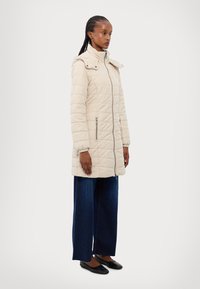Veste matelassée beige avec un col montant, fermeture �éclair frontale et deux poches latérales ; comprend une capuche amovible et un subtil détail de couture.