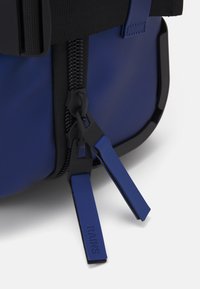Blå vanntett bag med teksturert overflate, utstyrt med svart hardware og glidelåser, samt blå gummihåndtak merket "RAINS."